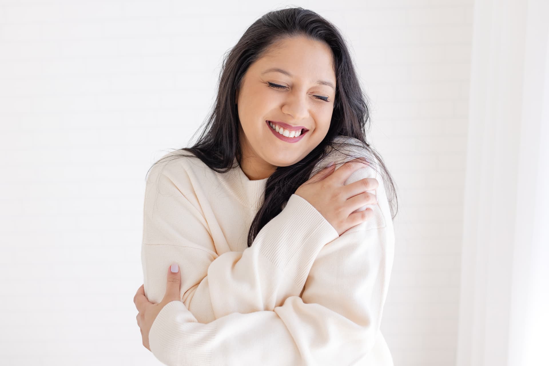 Tatiane Domingues de suéter claro, sorrindo, em um abraço acolhedor a si mesma